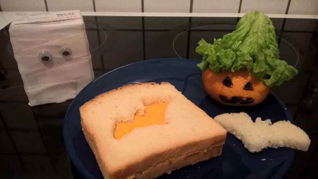 Auf dem Bild versammeln sich drei Halloween-Deko-Ideen. Eine bemalte Mandarine, ein ausgestochenes Brot und ein Milchpäckchen, das als Mumie verkleidet wurde. Wenn man auf das Bild klickt gelangt man zum eiweißarmen Rezept für Menschen mit PKU.