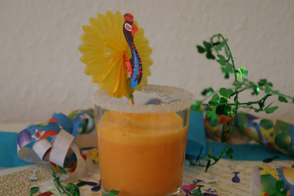 Ein Glas mit orangefarbenem Getränk. Das Glas hat einen Zuckerrand und im Getränk steckt ein Deko-Pfau. Silvesterdekoration im Hintergrund aus Luftschlangen und Kleeblättern. Wenn man auf das Bild klickt gelangt man zum eiweißarmen Rezept für Menschen mit PKU.