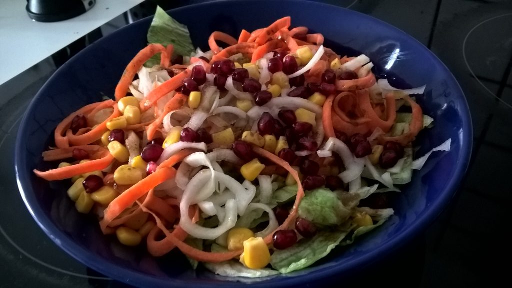 Ein Sommersalat in einem tiefen blauen Teller. Er besteht aus Gemüsespiralen, Mais, grünem Salat und Granatapfelkernen. Wenn man auf das Bild klickt gelangt man zum eiweißarmen Rezept für Menschen mit PKU.