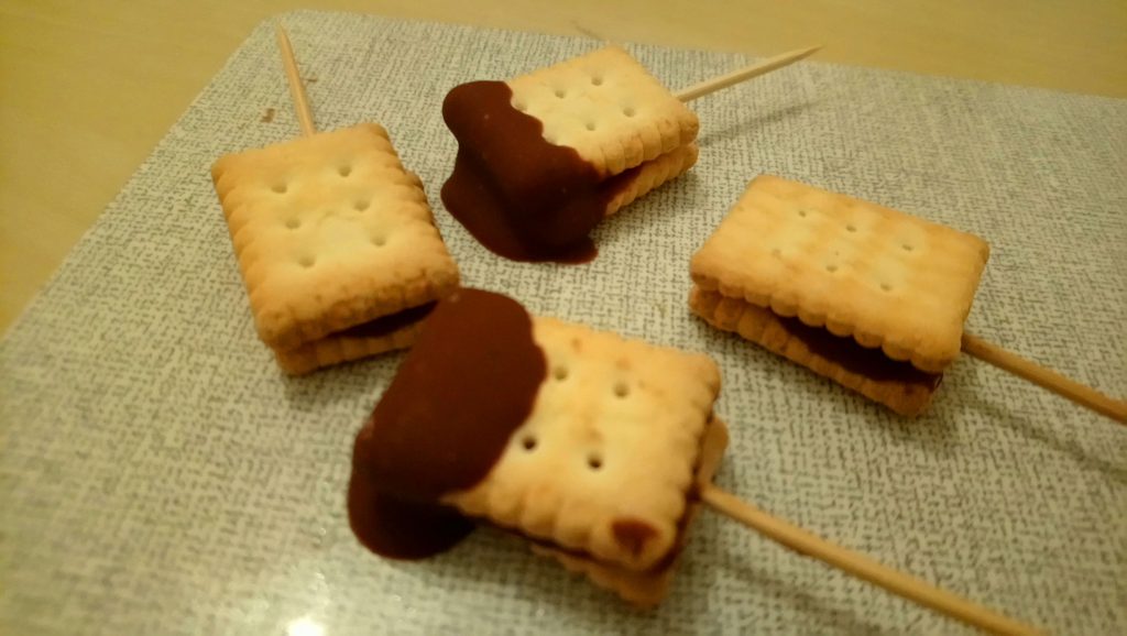 Auf einem Bretchen liegen vier kleine Kekse mit Schokolade, welche als 'am Stiel'-Lollis ausgestattet sind. Wenn man auf das Bild klickt gelangt man zum eiweißarmen Rezept für Menschen mit PKU.