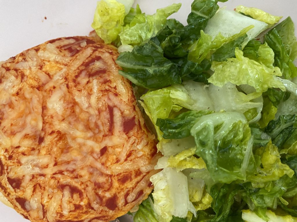 Ein eiweißarmes Pizza-Brötchen (Tomatensoße und veganer Käse als Belag) neben einer Portion grünem Blattsalat